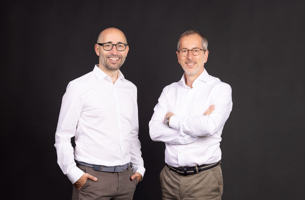 Zwei Männer in weißen Hemden und beigen Hosen stehen vor einem schwarzen Hintergrund. Der Mann auf der linken Seite trägt eine Brille, hat eine Glatze, lächelt und hat seine Hände in den Hosentaschen. Der Mann auf der rechten Seite hat graues Haar, trägt ebenfalls eine Brille, lächelt und hat die Arme verschränkt.