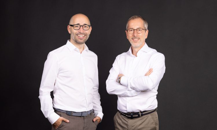 Zwei Männer in weißen Hemden und beigen Hosen stehen vor einem schwarzen Hintergrund. Der Mann auf der linken Seite trägt eine Brille, hat eine Glatze, lächelt und hat seine Hände in den Hosentaschen. Der Mann auf der rechten Seite hat graues Haar, trägt ebenfalls eine Brille, lächelt und hat die Arme verschränkt.